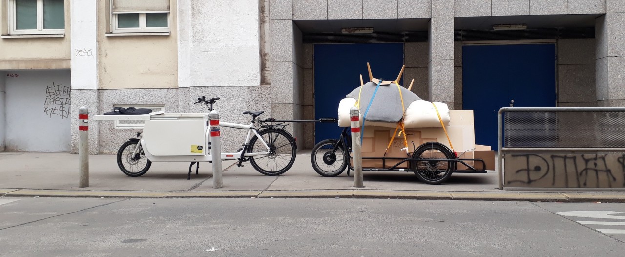 A side shot of a white cargo bike and black cargo trailer parked on a sidewalk, crammed with a pile of furniture which is topped by a bent mattress and an upside-down lounge chair, whose legs are pointing up and outwards like the spikes of a hedgehog
