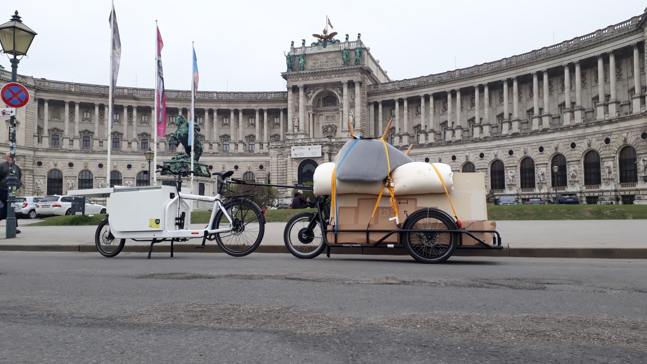 The bike, trailer and cargo from the last image, this time parked in front of the bent, scenic front of Vienna's Hofburg