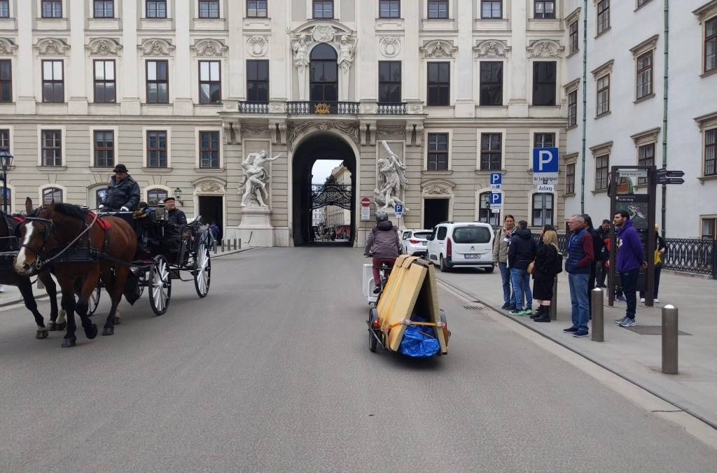 A street in a courtyard of Vienna's Hofburg, where a cargo bike with a trailer that is carrying two Tatami mats arranged like a triangle is passing by a horse carriage and a group of tourists