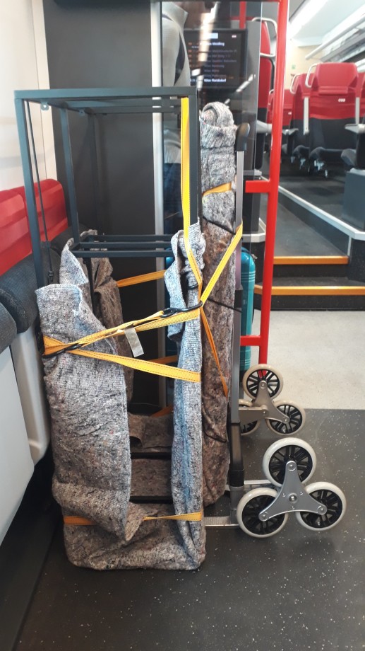 A hand truck carrying a cupboard, fixed with lashing straps and covered in a moving blanket, parked next to soft chairs on the S-Bahn