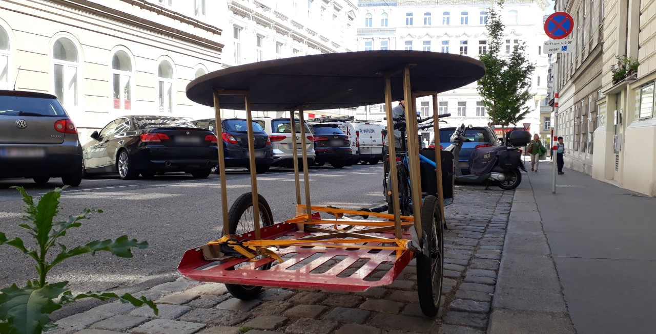A big round table fixed with lashing straps to a bright red cargo trailer parked next to a street lined with parked carss
